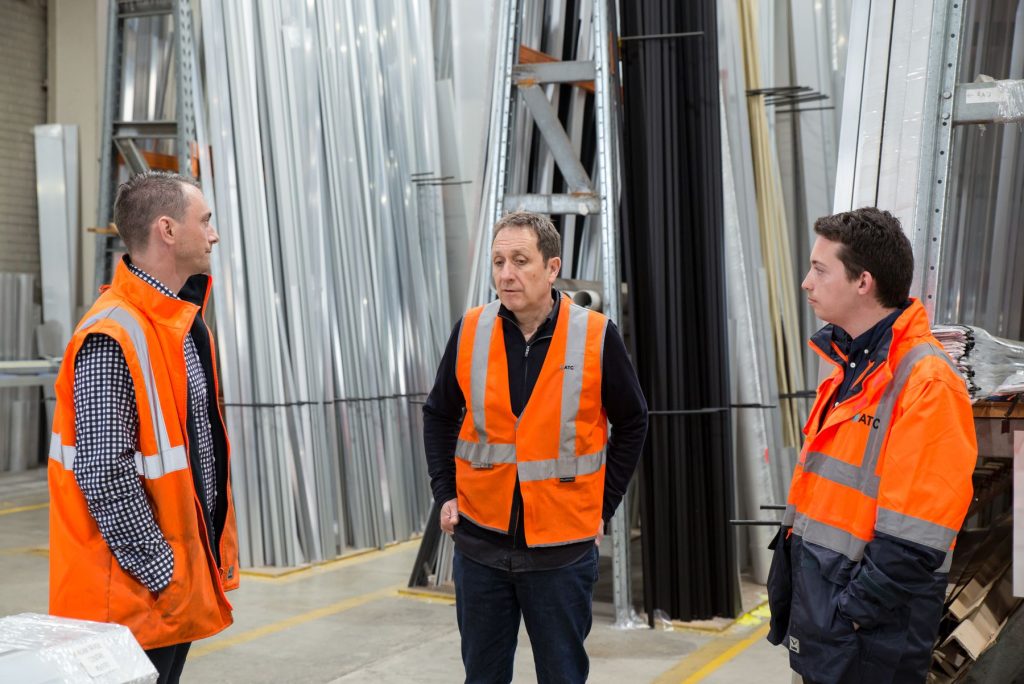 Tre lavoratori con gilet ad alta visibilita' parlano fra le scaffalature di profilati di un capannone industriale