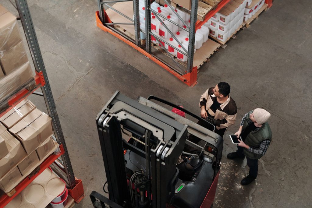 Veduta dall'alto di quattro persone che si coordinano accanto a un carrello elevatore fra le scaffalature di un magazzino