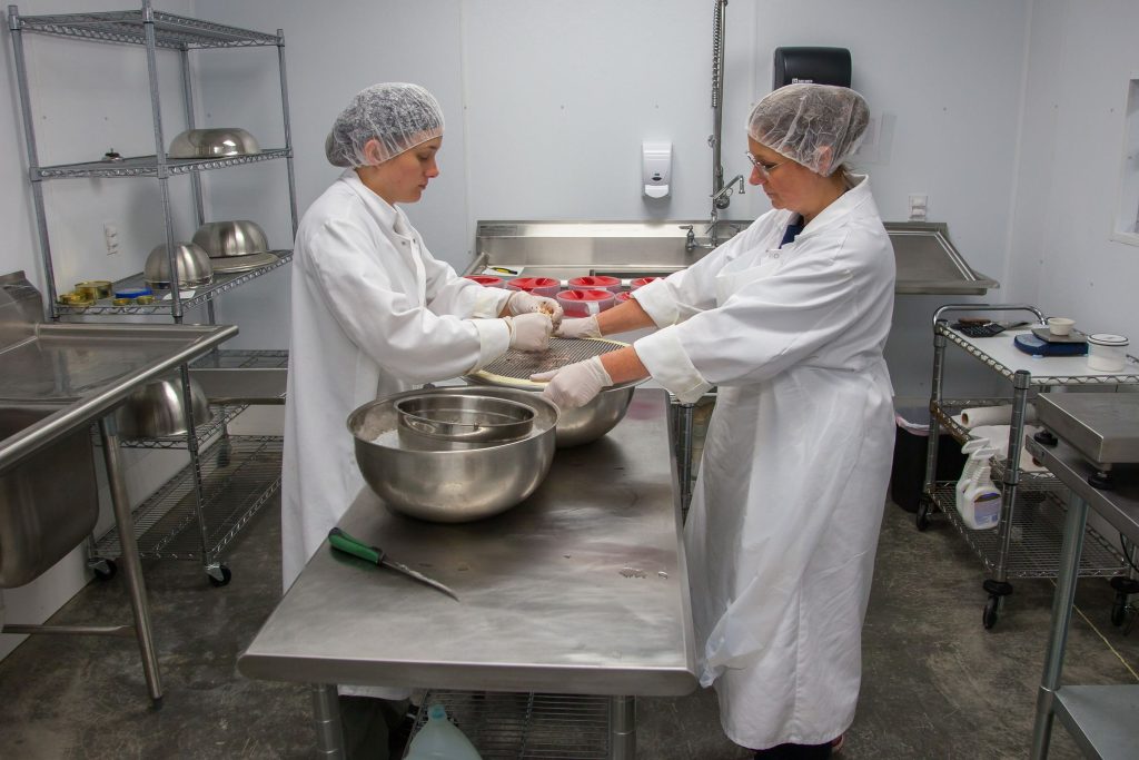 Due operatrici in camice, cuffia e guanti lavorano su un banco d'acciaio in una cucina professionale